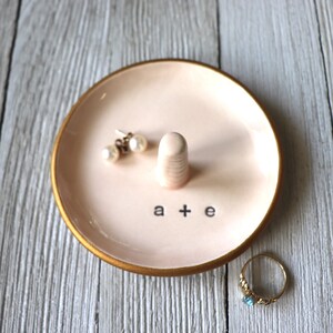 May include: A pink ceramic ring dish with a gold rim. The dish has a small, raised ring holder in the center and the letters "a + e" written on the bottom. Two pearl earrings are on the dish.