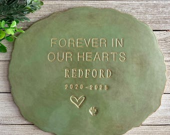 Puede incluir: Una lápida conmemorativa verde de forma irregular con las palabras "FOREVER IN OUR HEARTS" y el nombre "REDFORD" en relieve dorado. También se incluyen los años "2020-2026", un corazón y una huella de pata.