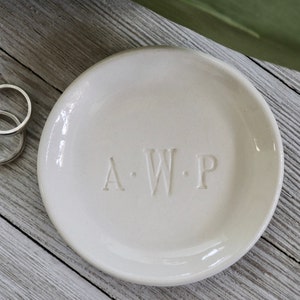 Puede incluir: Un plato redondo de cerámica blanquecina con las iniciales "A.W.P." grabadas en la superficie. El plato tiene un acabado brillante y probablemente se utiliza para guardar objetos pequeños. Dos anillos de plata son visibles en el lado izquierdo de la imagen.