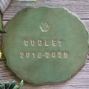 Pode incluir: Uma lápide redonda de cerâmica verde sálvia para animais de estimação. A pedra apresenta o nome "DUDLEY", os anos "2016-2025" e uma pegada. As bordas são irregulares, dando-lhe uma aparência rústica.