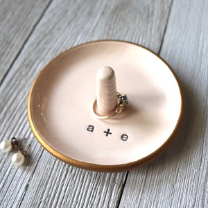 May include: A pink ceramic ring dish with a gold rim and a small white ceramic ring holder. The dish has the text "a + e" written on it.