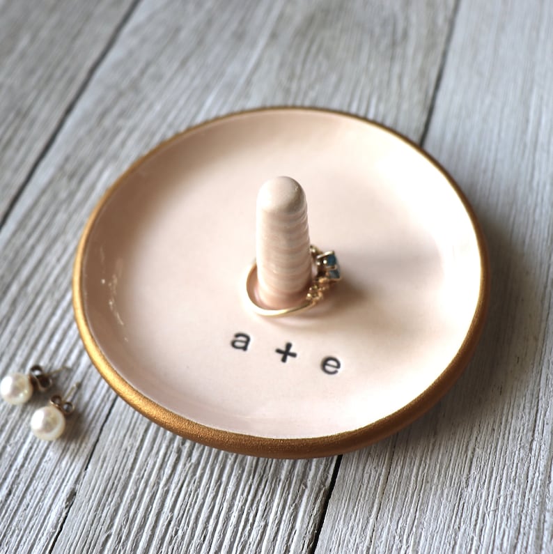 May include: A pink ceramic ring dish with a gold rim and a small white ceramic ring holder. The dish has the text "a + e" written on it.