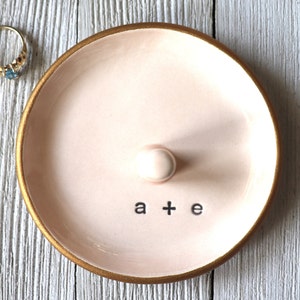 May include: A pink ceramic ring dish with a gold rim and a small knob in the center. The dish has the words "a + e" written on the bottom.