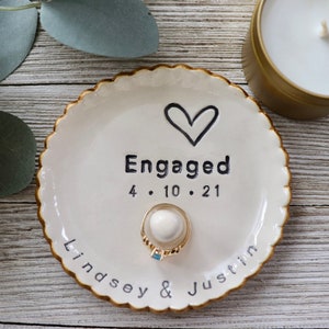 May include: White ceramic ring dish with a gold rim and a black heart design. The dish says "Engaged" with the date "4.10.21" and the names "Lindsey & Justin".
