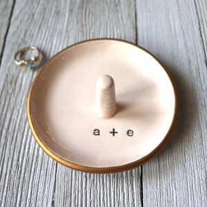 May include: A pink ceramic ring dish with a gold rim and a small white ceramic ring holder. The dish has the text "a + e" written on it.
