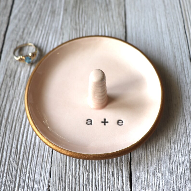 May include: A pink ceramic ring dish with a gold rim and a small white ceramic ring holder. The dish has the text "a + e" written on it.