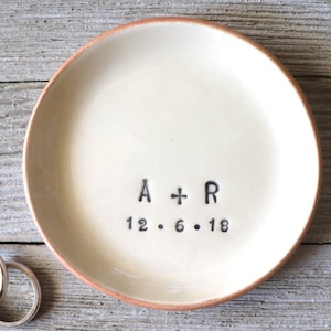 May include: A white ceramic ring dish with a brown rim. The dish has the text "A + R 12.6.18" inscribed on the bottom.