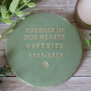 Op de afbeelding: Een ronde, saliegroene keramische gedenksteen met de woorden "FOREVER IN OUR HEARTS MAVERICK 2020-2025" en een pootafdruk. De steen heeft een licht onregelmatige rand en een goudkleurige rand.