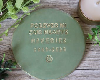 Puede incluir: Una piedra conmemorativa redonda de cerámica verde salvia con las palabras "FOREVER IN OUR HEARTS MAVERICK 2020-2025" y una huella de pata. La piedra tiene un borde ligeramente irregular y un borde dorado.
