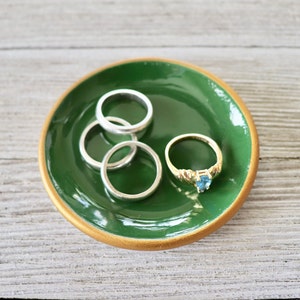 Op de afbeelding: Een groene keramische ringenschotel met een gouden rand. De schotel is gevuld met vier zilveren ringen en een gouden ring met een blauwe edelsteen.