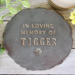 Może przedstawiać: Szara, kamienna tablica pamiątkowa z falistą krawędzią. Tekst "IN LOVING MEMORY OF TIGGER" jest wytłoczony w metalicznym kolorze, wraz z odciskiem łapy. Wzruszający hołd dla ukochanego zwierzaka.