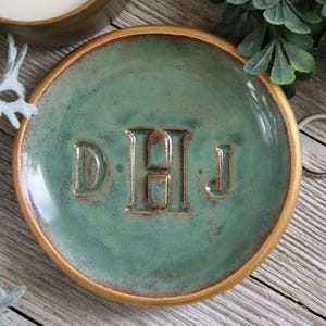 Puede incluir: Un plato para anillos de cerámica verde con un borde dorado y las iniciales "D.H.J" en una fuente serif.