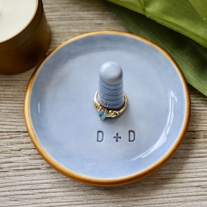 May include: A light blue ceramic ring dish with a gold rim and a small white ceramic ring holder. The dish has the letters "D + D" inscribed on the bottom.