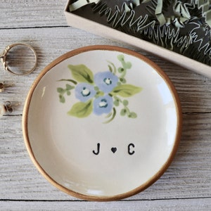 May include: A white ceramic ring dish with a gold rim. The dish is decorated with a blue floral design and the initials "J ♥ C".