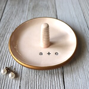 May include: A pink ceramic ring dish with a gold rim and a small white ceramic ring holder in the center. The dish has the words "a + e" written on it.