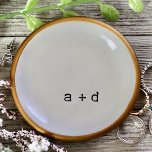 May include: A small white ceramic ring dish with a gold rim. The dish has the text "a + d" written in black on the bottom.