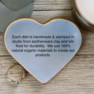 May include: A light blue heart-shaped ceramic dish with a gold rim. The dish is handmade and stamped in a studio from earthenware clay and kiln fired for durability. The text on the dish reads: "Each dish is handmade & stamped in studio from earthenware clay and kiln fired for durability. We use 100% natural organic materials to create our products."
