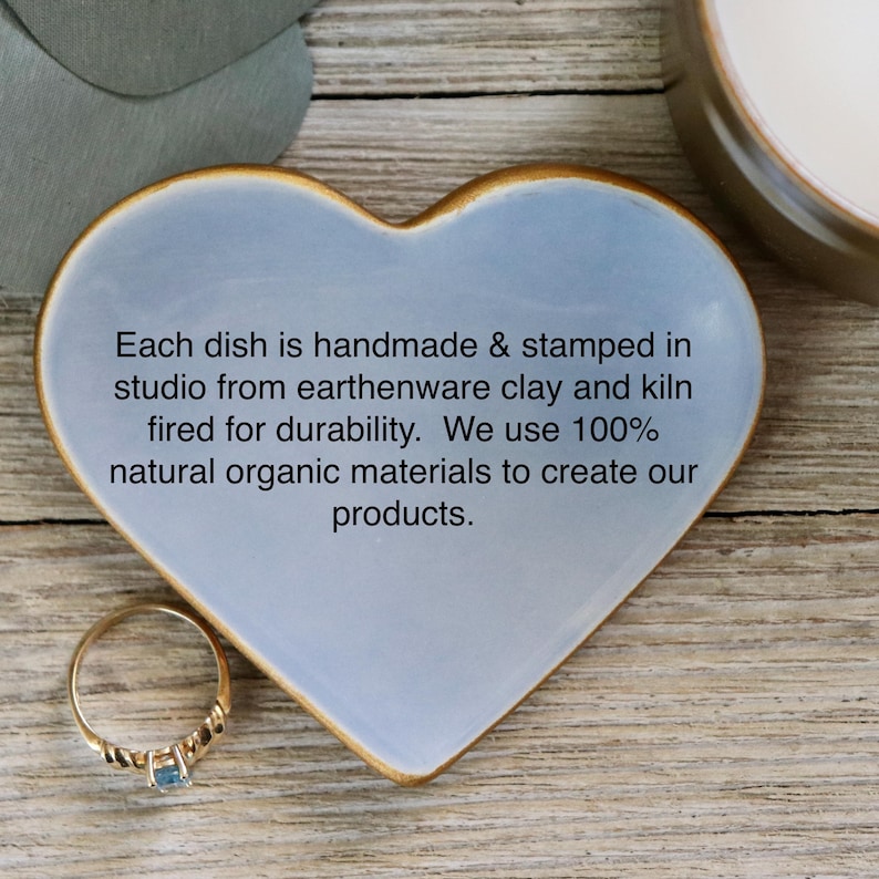 May include: A light blue heart-shaped ceramic dish with a gold rim. The dish is handmade and stamped in a studio from earthenware clay and kiln fired for durability. The text on the dish reads: "Each dish is handmade & stamped in studio from earthenware clay and kiln fired for durability. We use 100% natural organic materials to create our products."