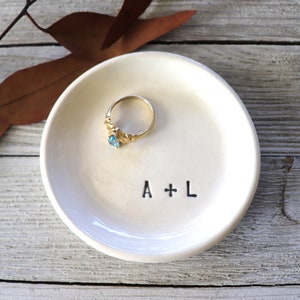 Puede incluir: Plato de cerámica blanco para anillos con las iniciales A + L grabadas en la parte inferior. Un anillo de oro con una piedra preciosa azul está sentado en el plato.
