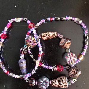 K&ouml;nnte beinhalten: Drei Armb&auml;nder mit Perlen in Lila, Rosa, Rot, Schwarz und Gold. Die Armb&auml;nder weisen verschiedene Perlenformen auf, darunter facettierte Kristalle, runde Perlen und gemusterte Fokusperlen. Die Armb&auml;nder sind auf einer dunklen Oberfl&auml;che angeordnet.