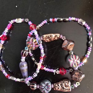 K&ouml;nnte beinhalten: Drei Armb&auml;nder mit Perlen in verschiedenen Lila-, Rosa-, Rot-, Schwarz- und Goldt&ouml;nen. Die Armb&auml;nder weisen verschiedene Perlenformen auf, darunter facettierte Kristalle, runde Perlen und gemusterte Fokusperlen. Die Armb&auml;nder liegen auf einer schwarzen Oberfl&auml;che.