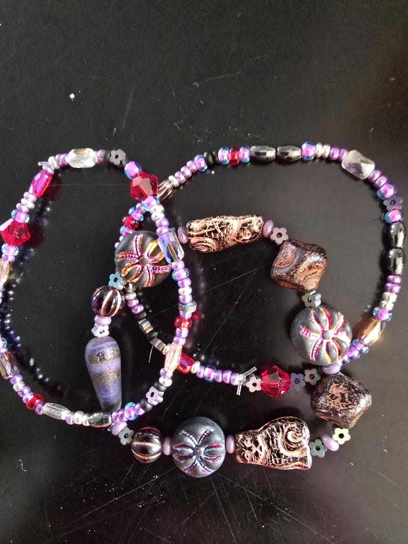 K&ouml;nnte beinhalten: Drei Armb&auml;nder mit Perlen in verschiedenen Lila-, Rosa-, Rot-, Schwarz- und Goldt&ouml;nen. Die Armb&auml;nder weisen verschiedene Perlenformen auf, darunter facettierte Kristalle, runde Perlen und gemusterte Fokusperlen. Die Armb&auml;nder liegen auf einer schwarzen Oberfl&auml;che.