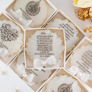Op de afbeelding: Een verzameling decoratieve onderzetters in witte dozen, elk met een uniek ontwerp en gouden rand. De onderzetters bevatten kalligrafie en de namen "HOUSSEM & AMINA" met de datum "25-12-2025". Witte linten en parels voegen een delicate touch toe.