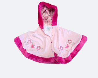 Puede incluir: Una capa con capucha de color rosa claro con un borde de piel sintética rosa intenso en la capucha y el dobladillo. La prenda incluye un cierre de botones frontal y motivos bordados de flores y mariposas en hilo rosa y blanco.