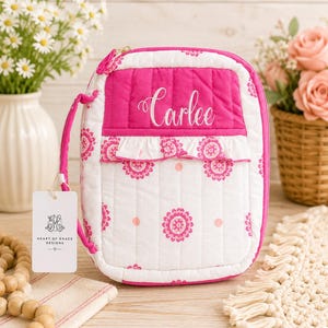 May include: A quilted fabric case with a zipper closure, featuring a white background with pink mandala-style patterns and a solid pink top section embroidered with the name Carlee in white script. A small white ruffled trim separates the sections.