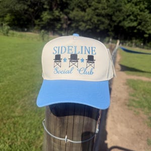 Op de afbeelding: Een crèmekleurige baseballpet met een lichtblauwe klep. Het voorpaneel is voorzien van geborduurde tekst SIDELINE Social Club in het blauw, vergezeld van drie zwarte klapstoelpictogrammen en twee lichtblauwe sterren tussen de stoelen.