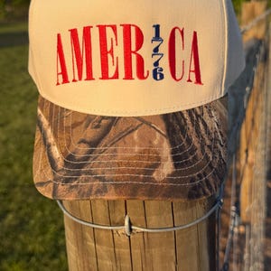 Könnte beinhalten: Beige und Camouflage Baseballkappe. Auf der Vorderseite der Kappe steht das Wort "AMERICA" in Rot, mit "1776" in Blau. Die Kappe liegt auf einem Holzpfosten.