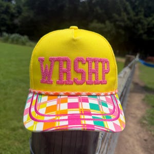 Könnte beinhalten: Eine leuchtend gelbe Trucker-Cap mit dem Schriftzug WRSHP, der in pinkfarbenen Blockbuchstaben auf die Vorderseite gestickt ist. Der Schirm zeigt ein buntes Karomuster in Pink, Orange und Grün, akzentuiert durch eine geflochtene Kordel.