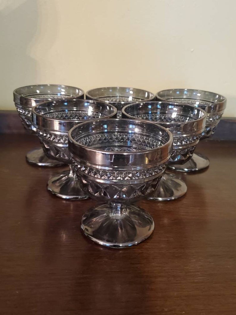 May include: Set of six silver-plated glass dessert cups with a diamond pattern. The cups have a silver rim and a clear glass base.
