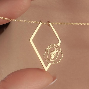 May include: A delicate gold necklace featuring a diamond-shaped pendant with a floral design. The pendant is suspended from a fine chain. The necklace is being held up by a hand.