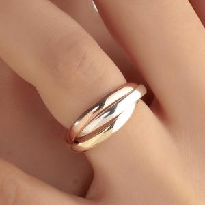 Peut inclure: Une bague dorée avec un design à trois bandes. La bague est portée au doigt, les mains au premier plan. Les bandes de la bague sont entrelacées, créant un look unique et élégant. L'arrière-plan est de couleur neutre.
