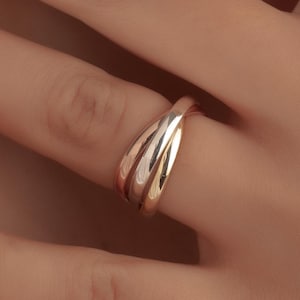 Könnte beinhalten: Ein dreifarbiges Ring mit ineinander verschlungenen Bändern aus Roségold, Gelbgold und Weißgold. Der Ring befindet sich an einem Finger, mit Hand und Arm im Hintergrund. Die Nägel sind mit French-Maniküre versehen.