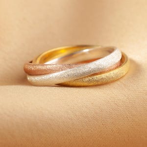 May include: Three interlocking rings in gold, rose gold, and silver tones rest on a beige fabric. The rings have a textured, matte finish, and the light reflects off the metal. The rings are a symbol of unity and commitment.