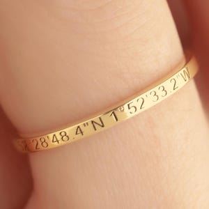 May include: A gold-colored ring with engraved coordinates, worn on a finger. The ring's surface is inscribed with the numbers and letters "28°48.4'N 1°52'33.2"W". The ring is a simple band design, and the background is a blurred skin tone.