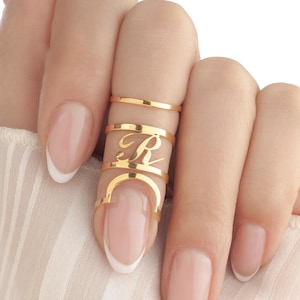 Könnte beinhalten: Eine Hand mit mehreren goldenen Ringen. Ein Ring ist ein schlichtes Band, ein anderer hat den Buchstaben "R" in Schreibschrift und ein dritter Ring hat ein gebogenes Design. Die Nägel sind mit einer French-Maniküre lackiert.