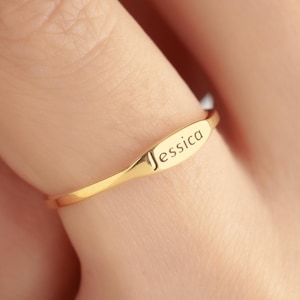 May include: A gold-colored ring with the name "Jessica" engraved on an oval-shaped plate. The ring is worn on a finger, showcasing the delicate design and personalized inscription. The ring is a piece of jewelry.