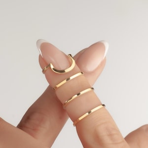 May include: A selection of gold-coloured rings. One ring is a curved shape, and the others are thin bands. The rings are worn on multiple fingers. The background is plain white.