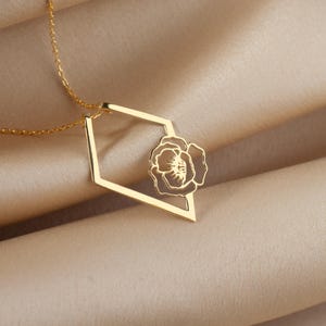 May include: A gold-colored necklace with a geometric pendant. The pendant features a flower design within a diamond-shaped frame. The necklace chain is delicate and the background is a soft, neutral fabric.