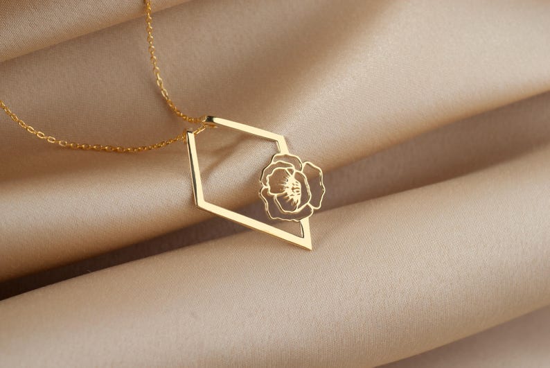 May include: A gold-colored necklace with a geometric pendant. The pendant features a flower design within a diamond-shaped frame. The necklace chain is delicate and the background is a soft, neutral fabric.