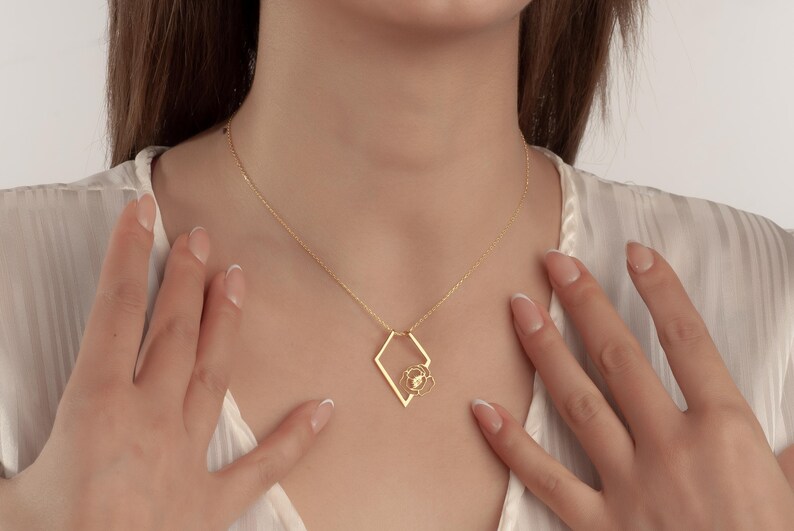 May include: A gold-colored necklace featuring a geometric pendant with a floral design. The pendant is suspended from a delicate chain. The necklace is worn against a white background.