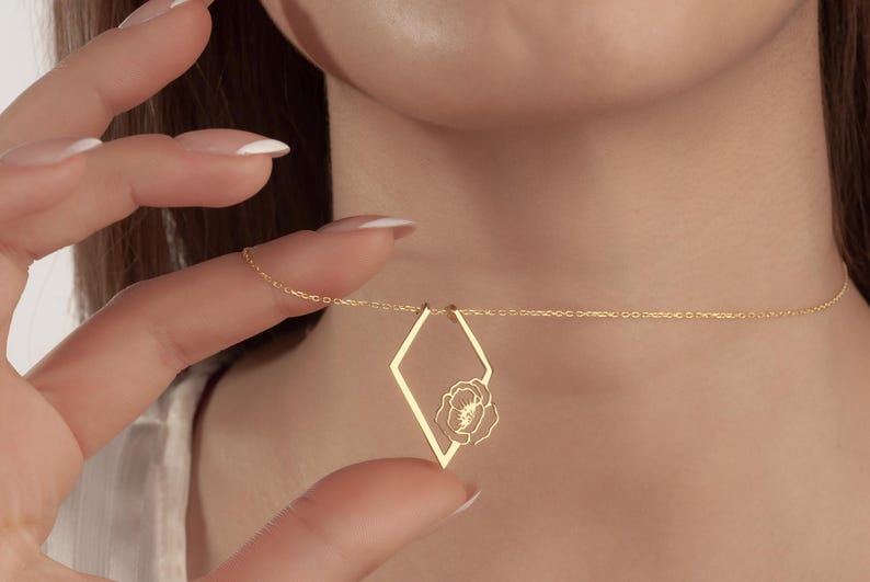 May include: A delicate gold necklace featuring a diamond-shaped pendant with a floral design. The pendant is suspended from a fine chain. The necklace is being held up by a hand.