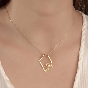May include: Gold-colored necklace featuring a geometric diamond-shaped pendant with a delicate rose design. The necklace is worn against a white, striped blouse. The chain is thin and the pendant is centered.