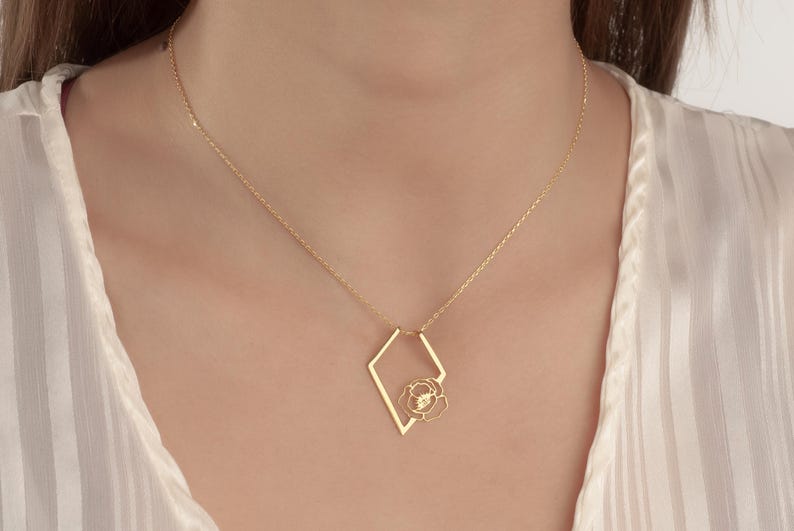 May include: Gold-colored necklace featuring a geometric diamond-shaped pendant with a delicate rose design. The necklace is worn against a white, striped blouse. The chain is thin and the pendant is centered.