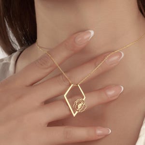 May include: A gold-colored necklace featuring a diamond-shaped pendant with a floral design. The delicate chain and pendant are visible against a light background. The necklace is a piece of jewelry.