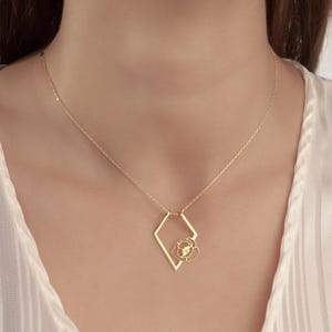 May include: A gold-colored necklace featuring a geometric diamond-shaped pendant with a delicate floral design at its center. The necklace hangs on a fine chain, creating an elegant and minimalist aesthetic. The jewelry is set against a soft, neutral background.