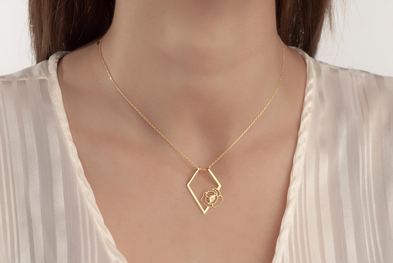May include: A gold-colored necklace featuring a geometric diamond-shaped pendant with a delicate floral design at its center. The necklace hangs on a fine chain, creating an elegant and minimalist aesthetic. The jewelry is set against a soft, neutral background.